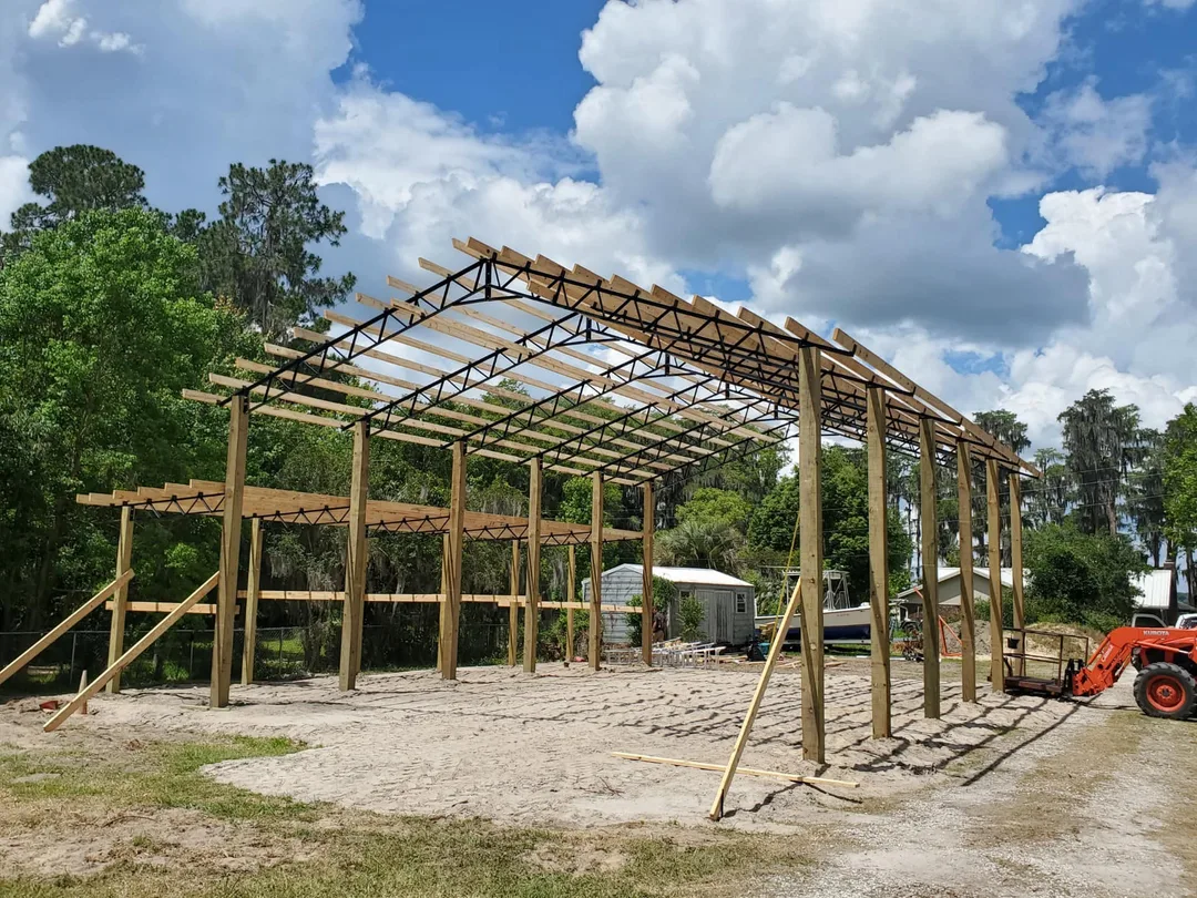 Pole barn steel-and-wood truss frame under construction on an open sand lot